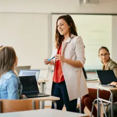 Nauczycielka z markerem wyjaśnia studentom, jak zostać nauczycielem po administracji. Uczniowie pracują na laptopach.
