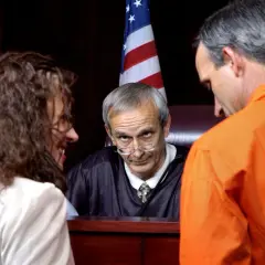 Sądowa scena: sędzia w todze patrzy na kobietę i mężczyznę w pomarańczowym stroju.