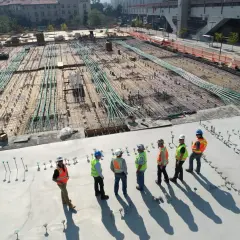 Grupa budowlańców w kamizelkach odblaskowych i kaskach omawia postęp prac na budowie. W tle widać zbrojenie i rury, a dalej budynki. Nie widać tu żadnego spychacza po angielsku.