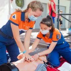 Dwie kobiety w strojach ratowniczych ćwiczą resuscytację na fantomie. Pokazują, kto może prowadzić kurs pierwszej pomocy.