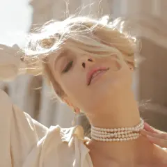 Kobieta z blond włosami, w białej bluzce, nosi naszyjnik z pereł. Pearl co to znaczy? To symbol elegancji i piękna.