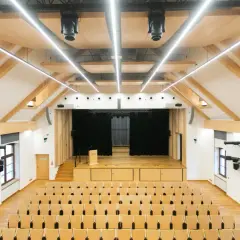 Nowa aula z drewnianymi elementami, sceną i rzędami krzeseł. Idealne miejsce na wykłady, koncerty i spotkania.