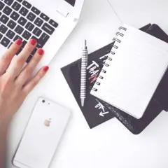 Dłoń z czerwonymi paznokciami pisze na laptopie. Obok leży telefon, notatnik i długopis. Co to znaczy? Praca, nauka, kreatywność.