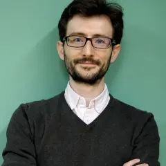 Uśmiechnięty nauczyciel po francusku w okularach i swetrze, z rękami skrzyżowanymi na piersi.