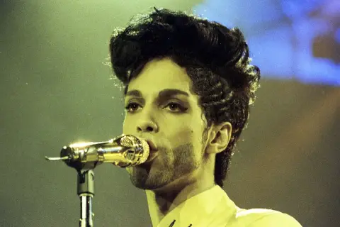 Prince, co to znaczy? Ikona muzyki śpiewa do złotego mikrofonu, jego charakterystyczna fryzura i makijaż podkreślają jego unikalny styl.