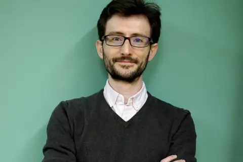 Uśmiechnięty nauczyciel po francusku w okularach i swetrze, z rękami skrzyżowanymi na piersi.