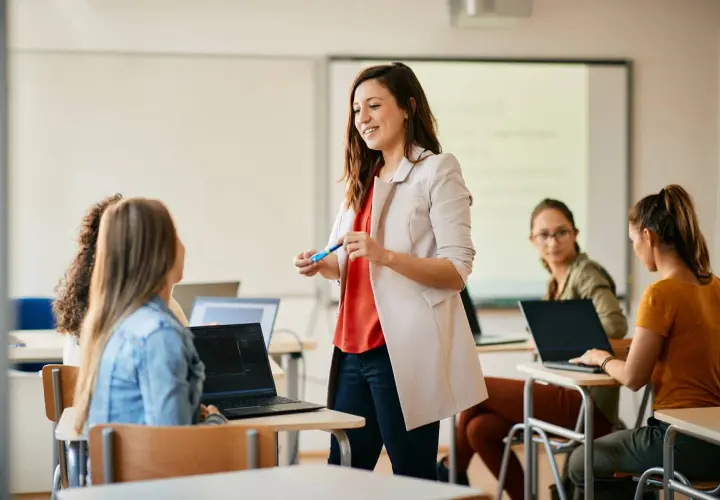 Nauczycielka z markerem wyjaśnia studentom, jak zostać nauczycielem po administracji. Uczniowie pracują na laptopach.