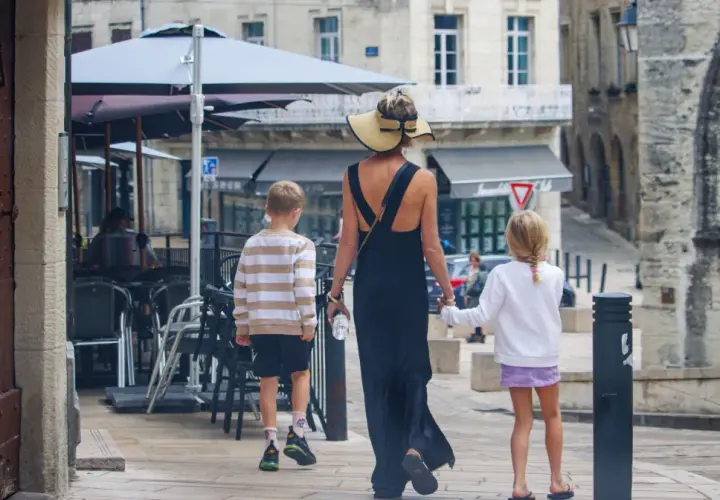 Kobieta w letniej sukience i kapeluszu prowadzi dwójkę dzieci, chłopca i dziewczynkę, ulicą. To francuskie dziecko, które zwiedza miasto.