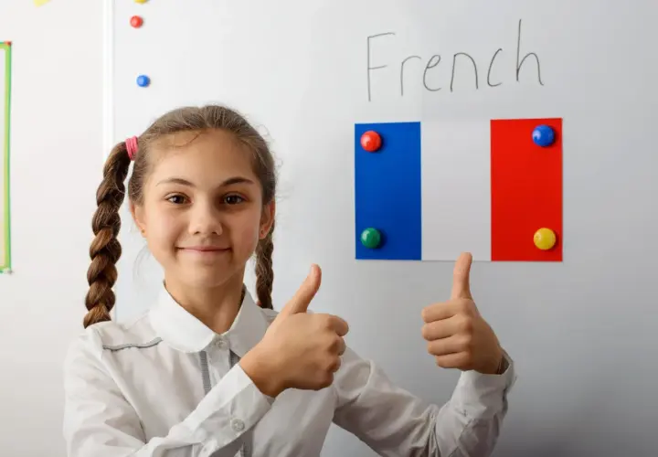 Uśmiechnięta dziewczynka z warkoczami pokazuje kciuki w górę. Na tablicy napis "French" i flaga Francji. Edukacja we Francji ciekawostki.