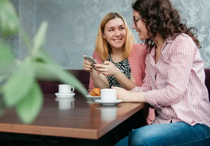 Dwie kobiety rozmawiają przy kawie, tworząc intymne tête-à-tête. Jedna pokazuje coś na telefonie, druga uśmiecha się.