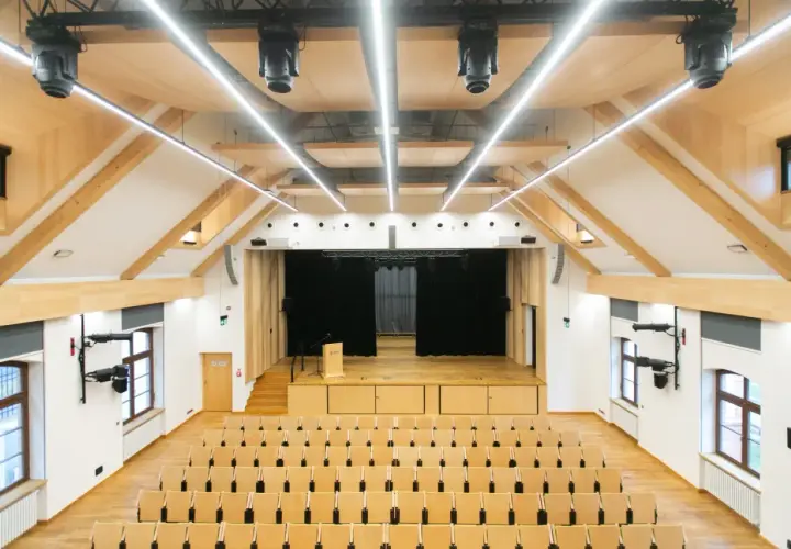 Nowa aula z drewnianymi elementami, sceną i rzędami krzeseł. Idealne miejsce na wykłady, koncerty i spotkania.