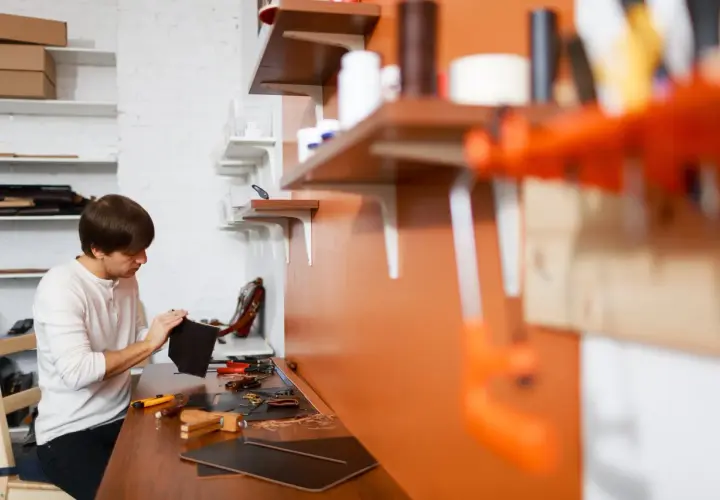 Mężczyzna pracuje w swoim atelier, tworząc skórzane wyroby. Na stole narzędzia i materiały.
