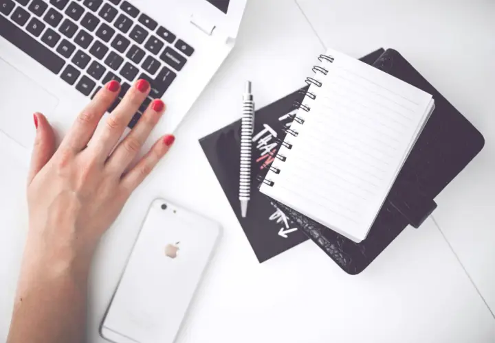 Dłoń z czerwonymi paznokciami pisze na laptopie. Obok leży telefon, notatnik i długopis. Co to znaczy? Praca, nauka, kreatywność.