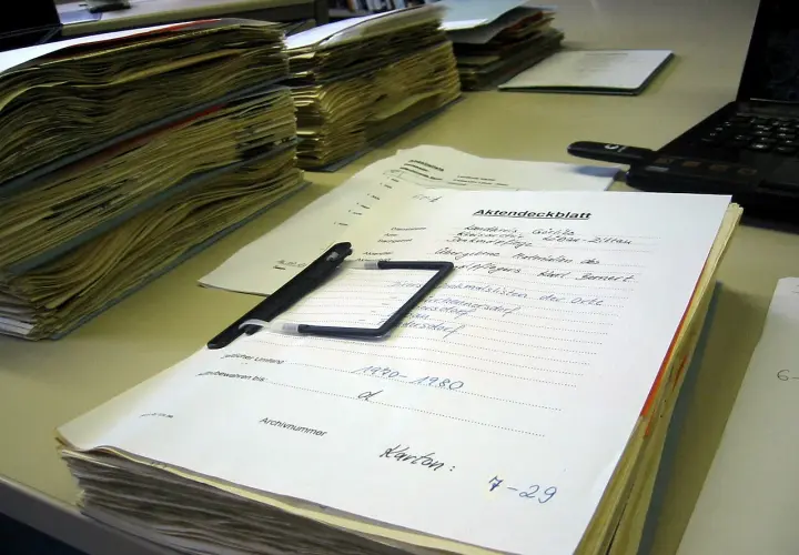 Stosy dokumentów, długopis i laptop na biurku. Na jednym z dokumentów widnieje napis "Akten-dossier co to", sugerujący porządkowanie akt.