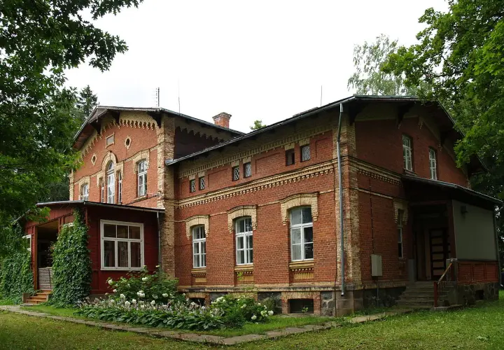 Stary, ceglany maison otoczony zielenią, z werandą i kwitnącymi krzewami.