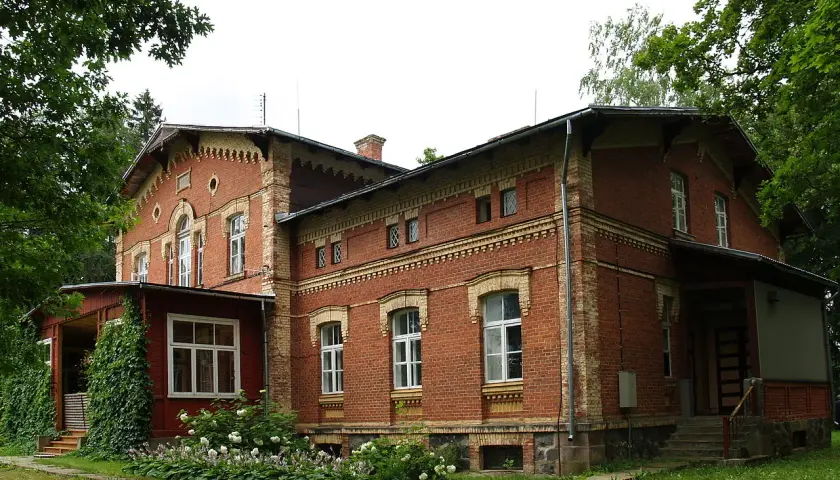 Stary, ceglany maison otoczony zielenią, z werandą i kwitnącymi krzewami.