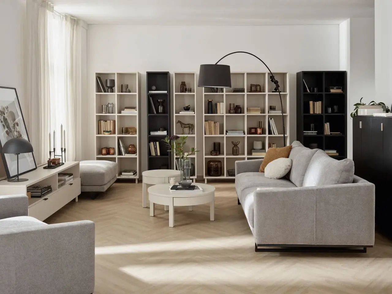 Nowoczesny living room z szarą sofą, regałami pełnymi książek i dekoracji, okrągłym stolikiem kawowym i lampą łukową.