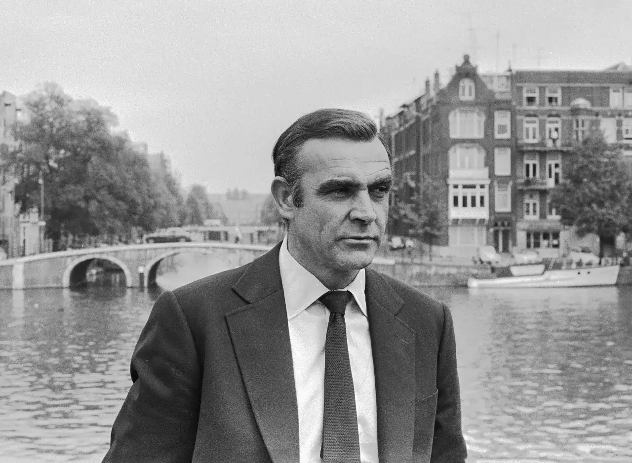 Sean Connery, co oznacza sir, w garniturze na tle kanału i kamienic w Amsterdamie.