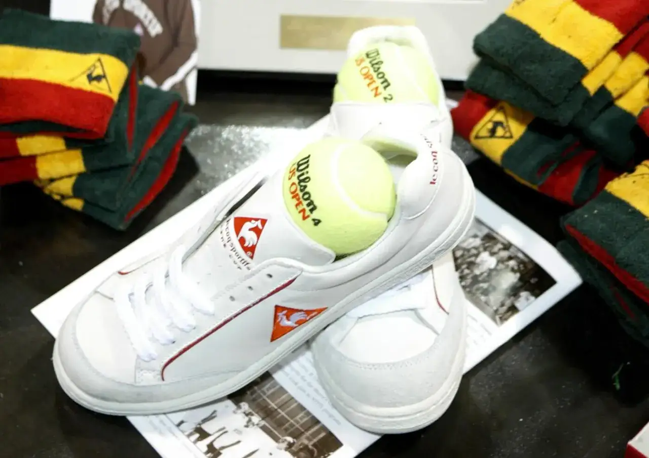 Białe buty tenisowe Le Coq Sportif z piłkami Wilson US Open. Obok stosy ręczników w kolorach francuskiej flagi.