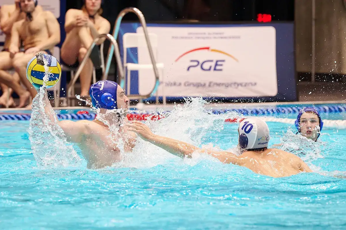 Zacięta walka w wodzie podczas meczu piłki wodnej. Gracze w czepkach walczą o piłkę, tryska woda. To emocjonujące piscine po polsku.