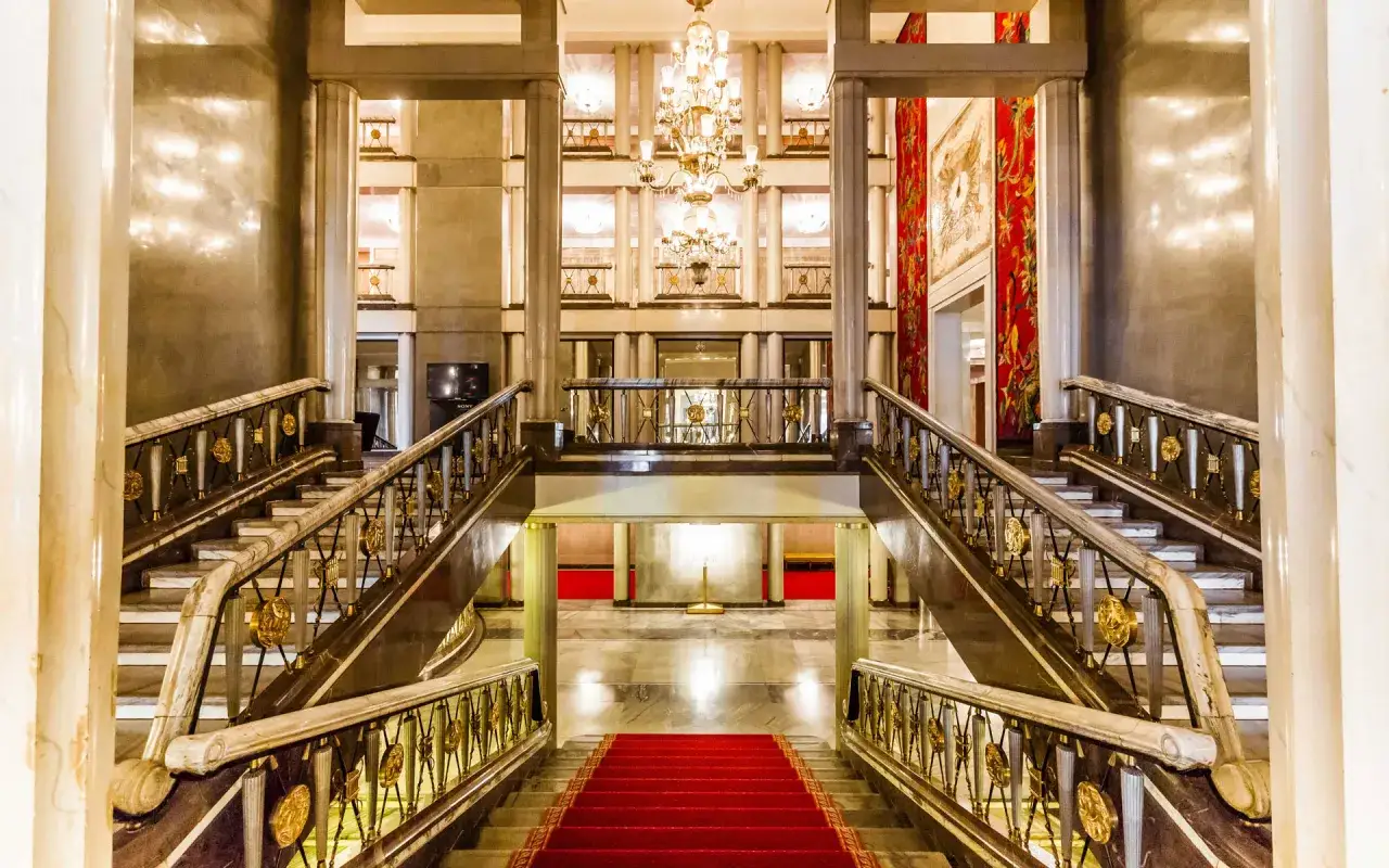 Elegancki, marmurowy foyer z dwoma biegnącymi schodami, ozdobionymi złotymi detalami i czerwonym dywanem.
