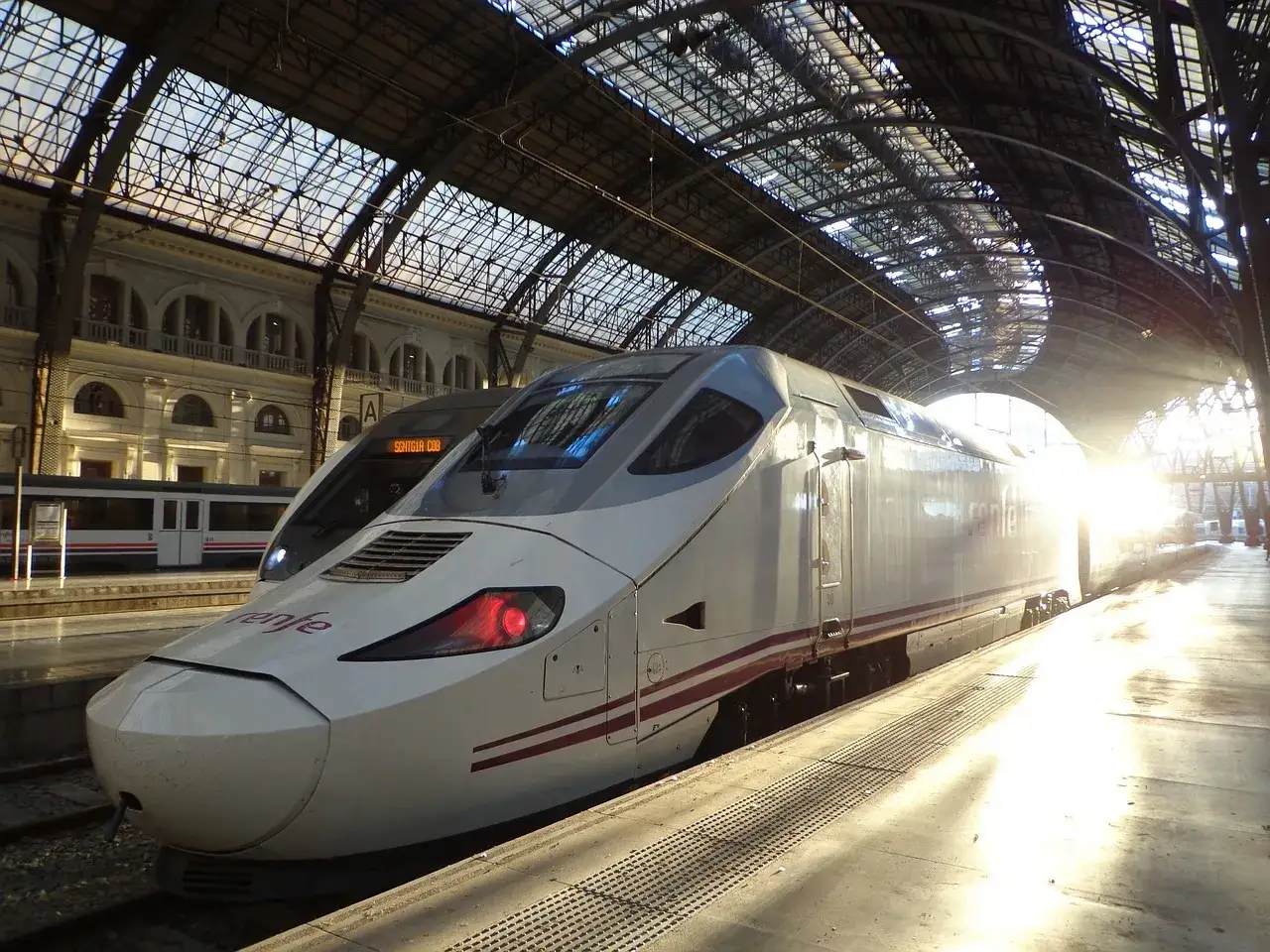 Nowoczesne pociągi Renfe na dworcu pod szklanym dachem. Słońce przebija się przez konstrukcję, tworząc refleksy.