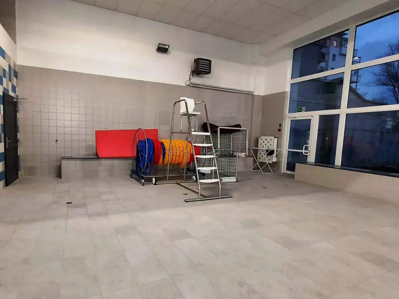 Puste wnętrze basenu, gotowe na zajęcia. Widać drabinkę, czerwony materac i zwinięte boje.