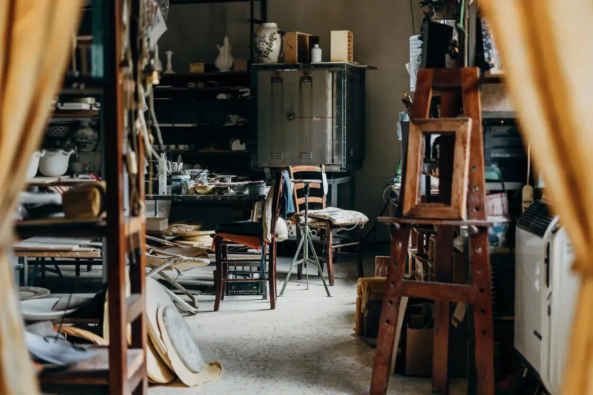 Wnętrze atelier artystycznego. Widać tu piec, sztalugi, krzesła, półki z naczyniami i materiałami do tworzenia.