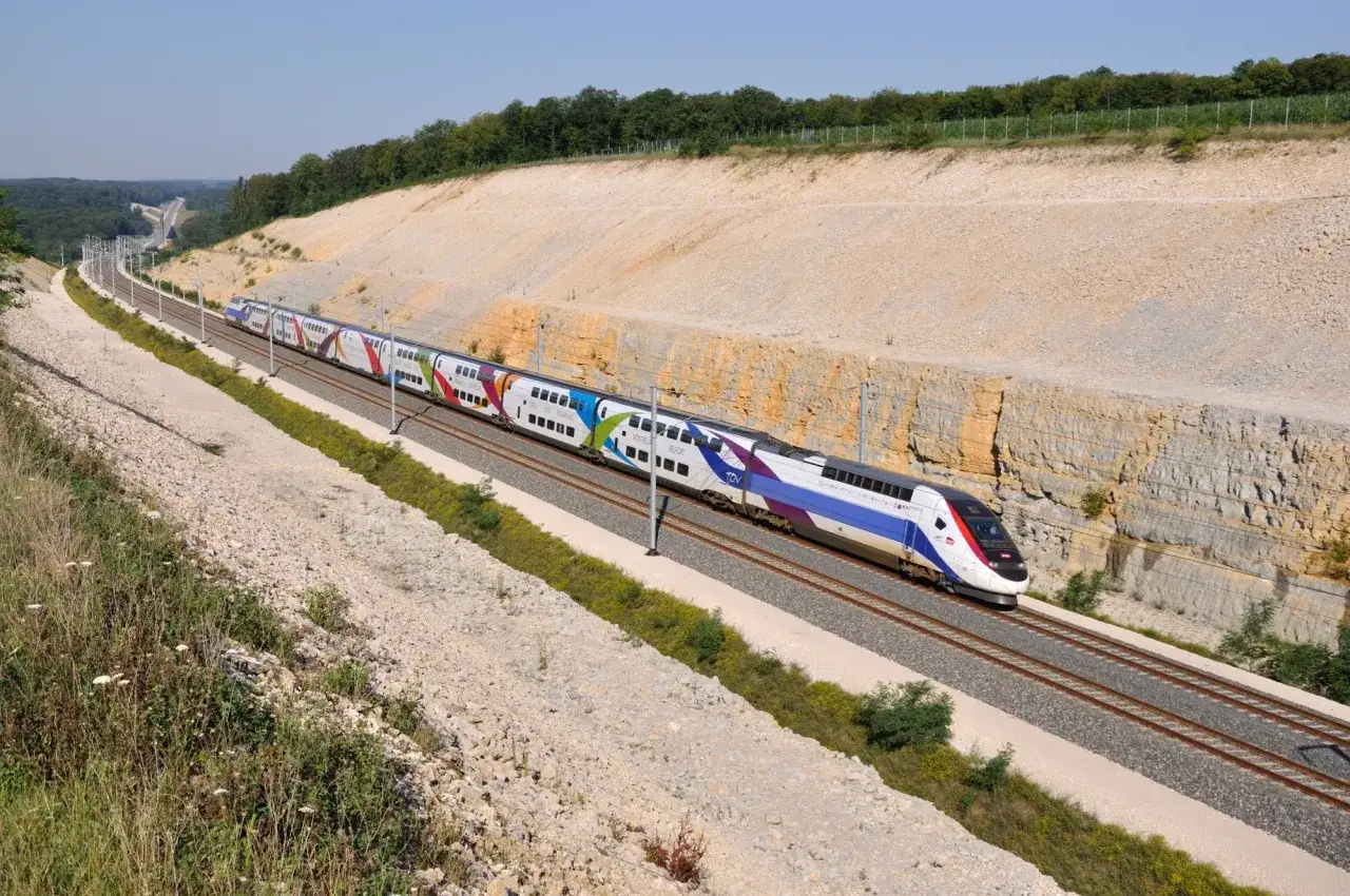 Kolorowy pociąg TGV mknie po torach wciętych w skarpę. Te ze we krajobrazu tworzą malowniczą scenerię.