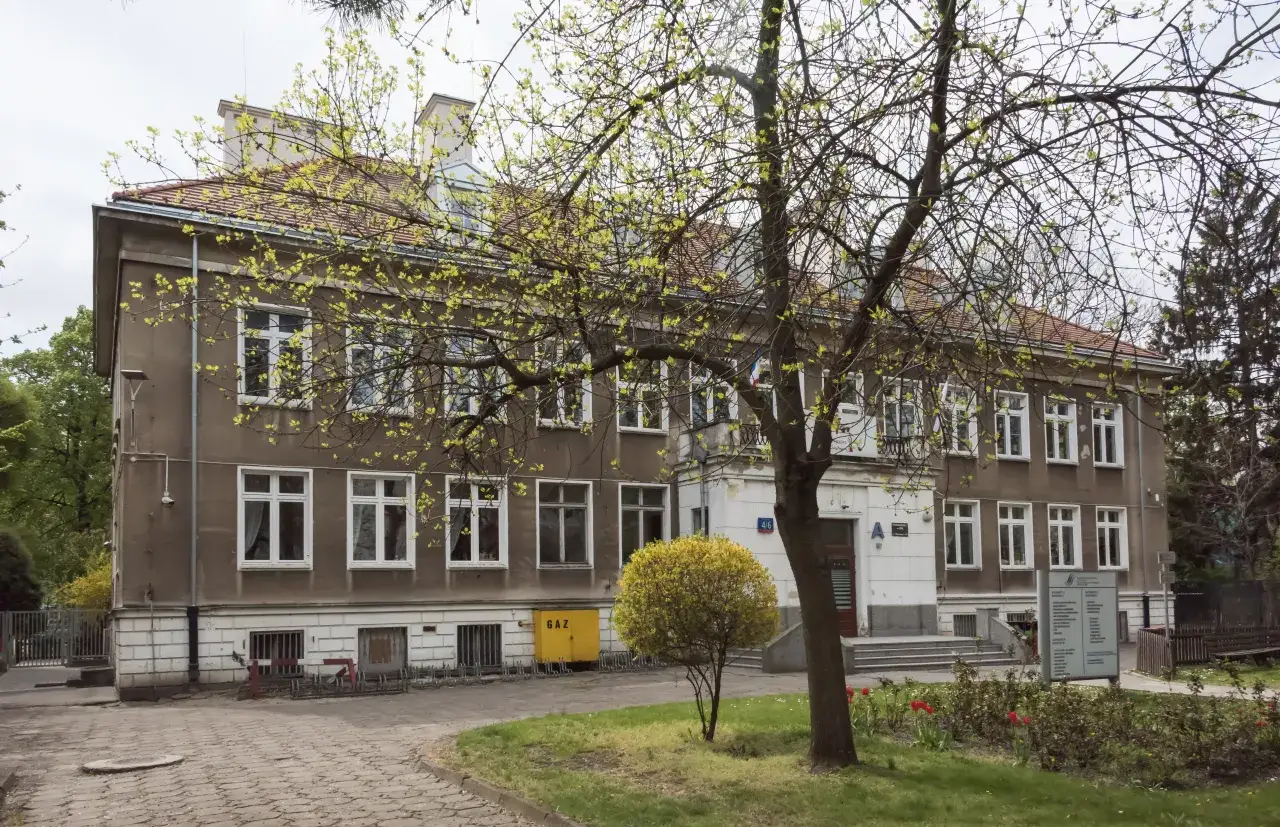 Budynek z żółtą skrzynią gazową i tablicą informacyjną. Wiosna, młode liście na drzewach. To może być szkoła po francusku.
